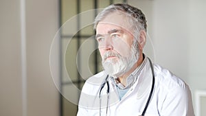 Thoughtful sad senior doctor looking away indoors. Portrait of grey-haired Caucasian man looking at camera and crossing