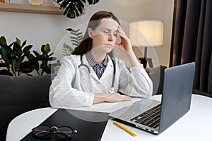 Thoughtful sad young woman doctor sit at work desk with pc laptop, pondering problem solution, pensive therapist physician wearing