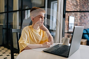 Thoughtful schoolboy typing on laptop computer and thinking with hands on chin. Pupil child boy learning lesson at