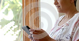 Thoughtful senior biracial woman using smartphone at window at home, slow motion