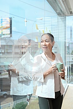 Thoughtful senior female business leader holding coffee cup and standing near large window at office with city buildings