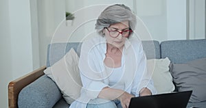 Thoughtful senior woman freelancer wearing eyeglasses typing on laptop sitting sofa.