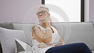 Thoughtful senior woman with grey hair sitting on the living room sofa at home, showing a serious expression, reflecting on a