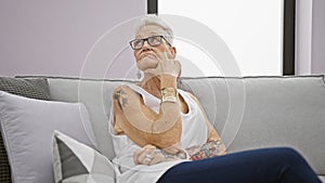 Thoughtful senior woman with grey hair sitting on the living room sofa at home, showing a serious expression, reflecting on a