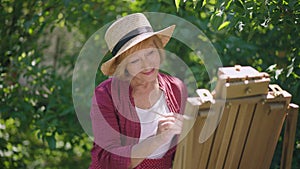 Thoughtful senior woman painting in sunshine outdoors and smiling looking at camera. Portrait of confident talented