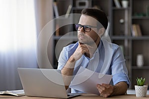 Thoughtful serious businessman in glasses touching chin, holding document