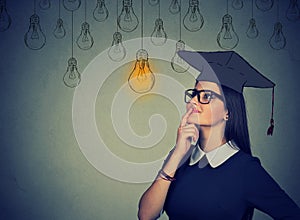 Thoughtful student woman in cap gown looking up at light bulb thinking