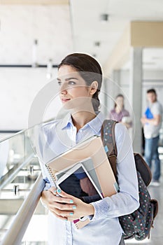 Thoughtful teenage girl with books and digital tablet standing in university corridor