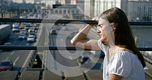 Thoughtful teenage girl is watching moving cars over road sitting on bridge in city in summer