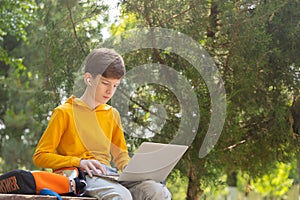 Thoughtful teenager boy resting. Holding and using a laptop for networking on a sunny day, outdoors.