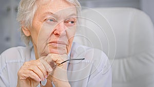 Thoughtful wise aged lady with sad eyes wrinkly face and short grey hair looks aside holding glasses in hand