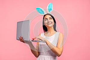 Thoughtful woman in bunny ears holding laptop, video call, shopping online in studio on pink background, easter