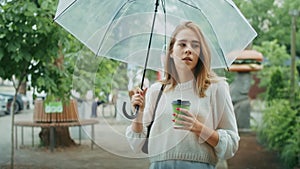 Thoughtful woman holding umbrella and coffee cup on a city street. Contemplative urban scene with lush greenery. Modern