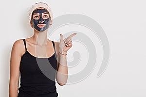 Thoughtful woman looks pensively up, applies black mask on face, wears hairband and sleeveless t shirt, points away at empty space