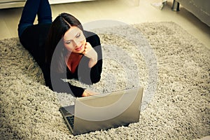 Thoughtful woman lying on the carpet with laptop