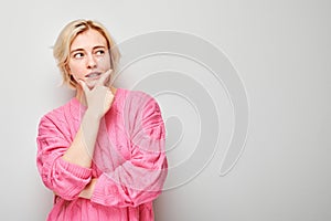 Thoughtful woman in pink blouse with hand on chin, looking away on a gray background