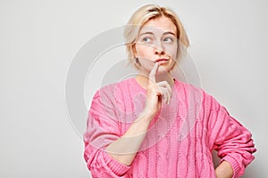 Thoughtful woman in pink sweater with finger on lips, looking away, on light background