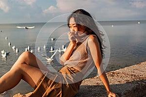 Thoughtful woman sitting on the sandy shore looking at a lake and cloudy sky with flying seagulls.