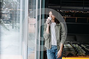 Thoughtful woman standing in a bedroom next to a window, drinking coffee