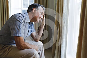 Thoughtful worried senior caucasian man sitting on sofa and looking through window