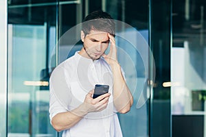 Thoughtful and worried young man, businessman, freelancer, businessman looks at the phone, holds his head, received bad news