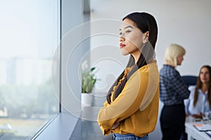 Thoughtful young Asian businesswoman looking through window at office