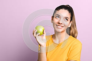 Thoughtful young attractive woman in yellow t-shirt holding green apple and smiling thinking about junk food