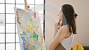 Thoughtful young beautiful hispanic woman artist, concentrating and doubting with the brush in hand, looking at her drawing on the