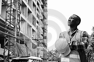 Thoughtful young black African man construction worker holding clipboard and hard hat with arms crossed at building site