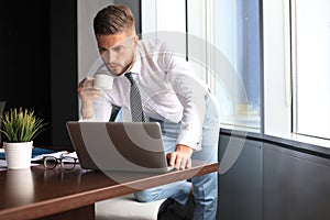 Thoughtful young business man is using computer and is drinking coffee while working in the office