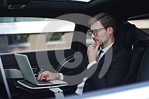 Thoughtful young businessman keeping hand on chin while sitting in the lux car and using his laptop