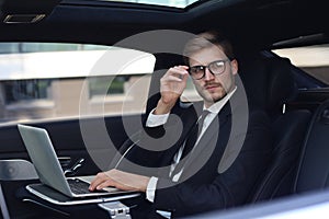 Thoughtful young businessman keeping hand on glasses while sitting in the lux car and using his laptop