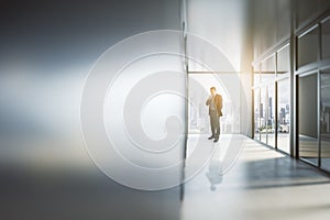 Thoughtful young businessman looking out of window in abstract bright light office interior with mock up place on silver wall.