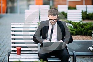 Thoughtful young businessman in suit sitting on wooden bench with coffee cup in hand and laptop placed on his lap.