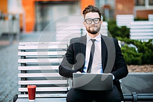 Thoughtful young businessman in suit sitting on wooden bench with coffee cup in hand and laptop placed on his lap.