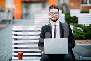 Thoughtful young businessman in suit sitting on wooden bench with coffee cup in hand and laptop placed on his lap.