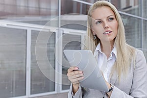 Thoughtful young businesswoman using digital tablet while looking away against office building