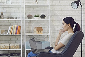 Thoughtful young businesswoman using laptop computer from home. Side view of freelancer working on new project indoors