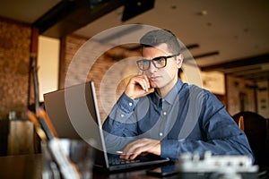 Thoughtful young caucasian businessman in glasses working on laptop computer. Pensive attractive hipster freelancer