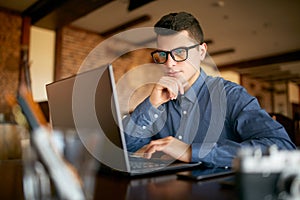 Thoughtful young caucasian businessman in glasses working on laptop computer. Pensive attractive hipster freelancer