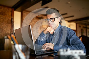 Thoughtful young caucasian businessman in glasses working on laptop computer. Pensive attractive hipster freelancer