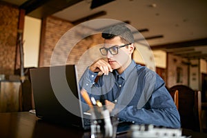 Thoughtful young caucasian businessman in glasses working on laptop computer. Pensive attractive hipster freelancer