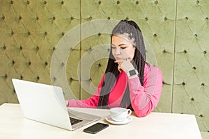 Thoughtful young freelancer with black dreadlocks in pink blouse is sitting in cafe and working on laptop, wait for new idea and