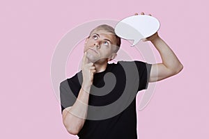 Thoughtful young man holding speech bubble while looking up over pink background