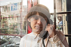 Thoughtful young man in light striped shirt makes call from old public telephone booth, city reflections in background