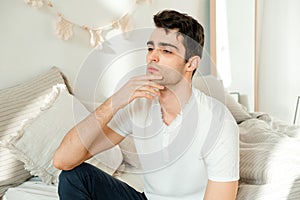 Thoughtful young man sitting on the bed in the bedroom, touching his chin and thinking. Thoughtful mature man resting on