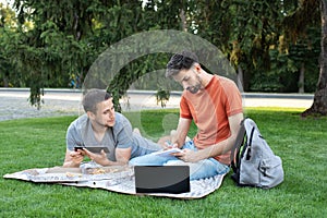 Thoughtful young man is sitting with a laptop on grass on a college campus and taking notes in notebook. Man explaining something