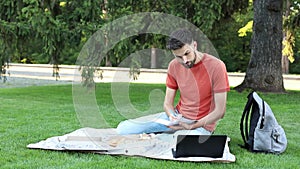 Thoughtful young man is sitting with a laptop on grass on a college campus and taking notes in notebook. Student learns during a b