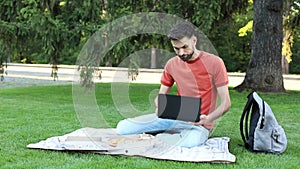 Thoughtful young man is sitting with a laptop on grass on a college campus and taking notes in notebook. Student learns during a b