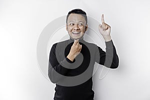 A thoughtful young man wearing a black shirt while looking and pointing at copy space above him, isolated by white background